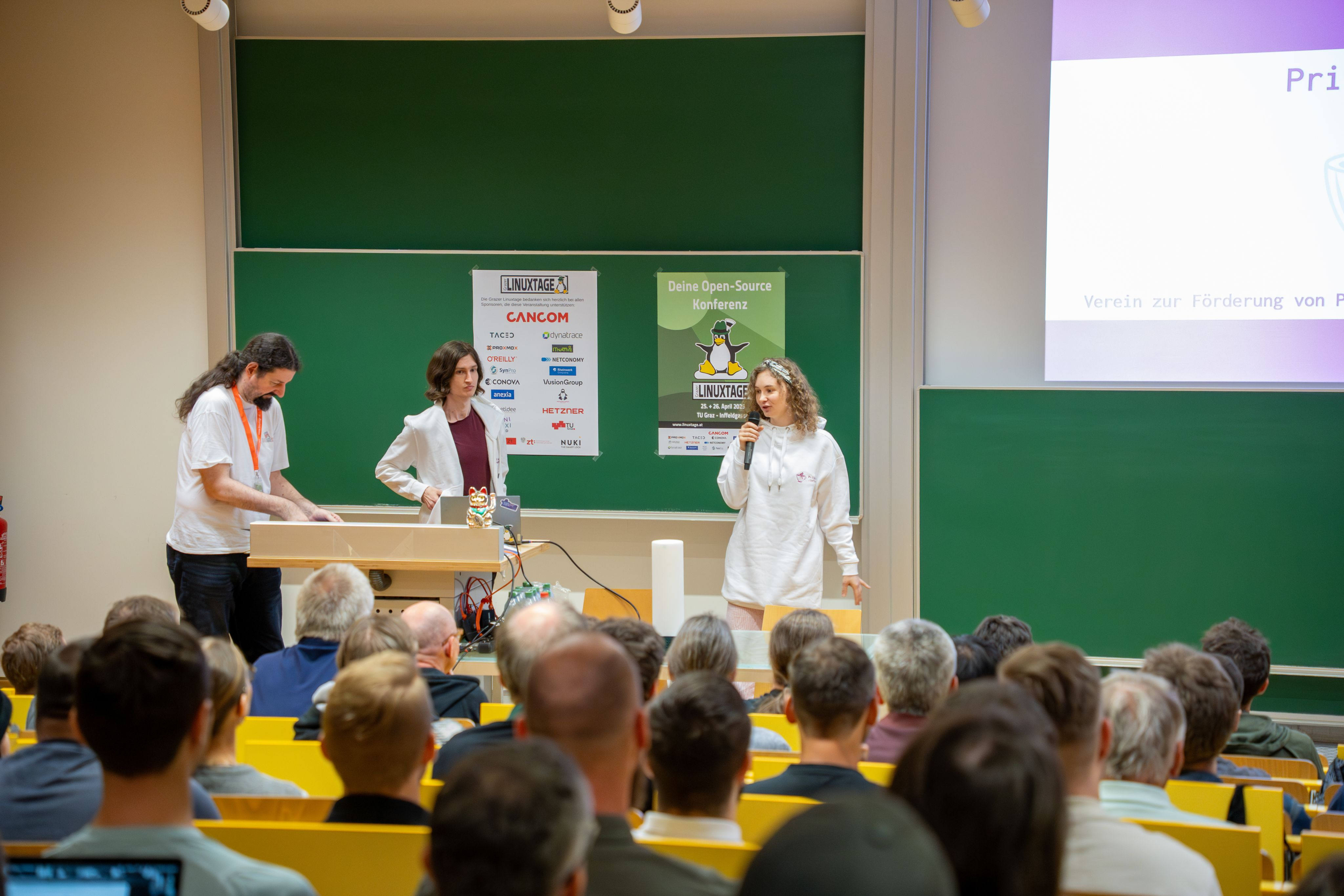Jupfi at the lightning talk. A  crowd is visible watching jupfi giving the lightning talk. The Linuxtage mascot (a penguin with a Steirerhut) is visible on a chalkboard.