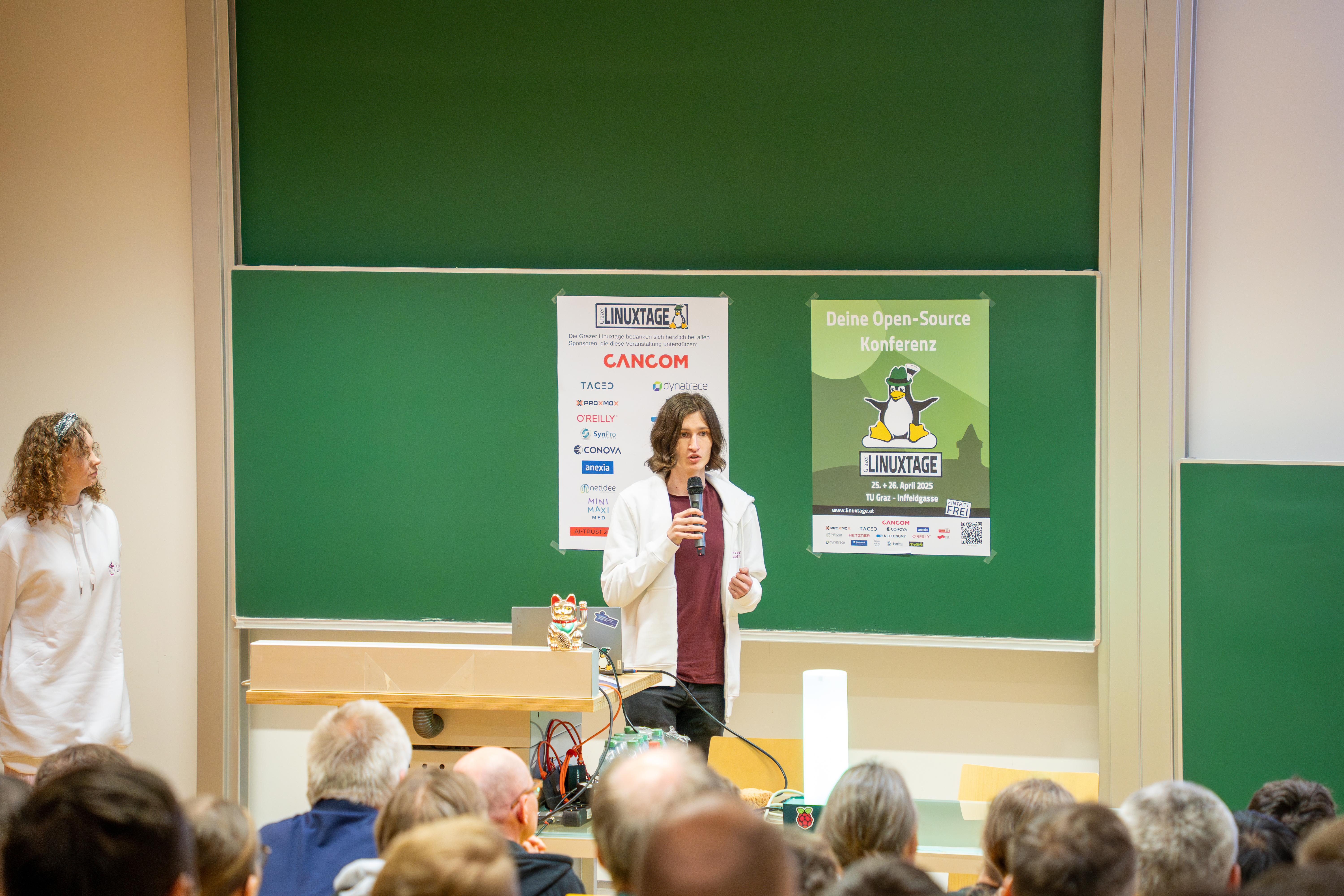 Kumi at the lightning talk. A  crowd is visible watching Kumi giving the lightning talk. The Linuxtage mascot (a penguin with a Steirerhut) is visible on a chalkboard.